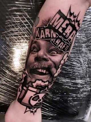 Detailed portrait tattoo on forearm featuring an expressive face with text banners above and below, rendered in black ink with shading.