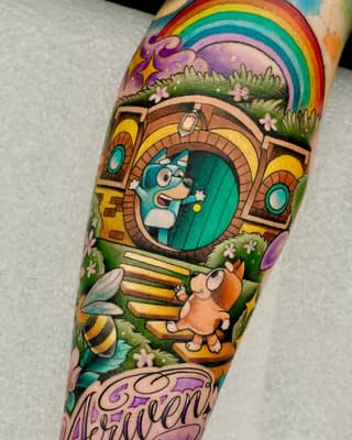 Colorful arm tattoo featuring a whimsical character in a round doorway, rainbow, mushrooms, small creatures, and green grass landscape with purple text below.