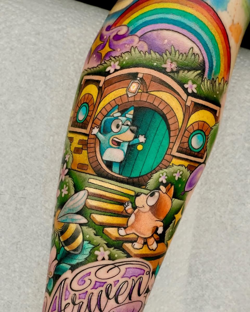 Colorful arm tattoo featuring a whimsical character in a round doorway, rainbow, mushrooms, small creatures, and green grass landscape with purple text below.