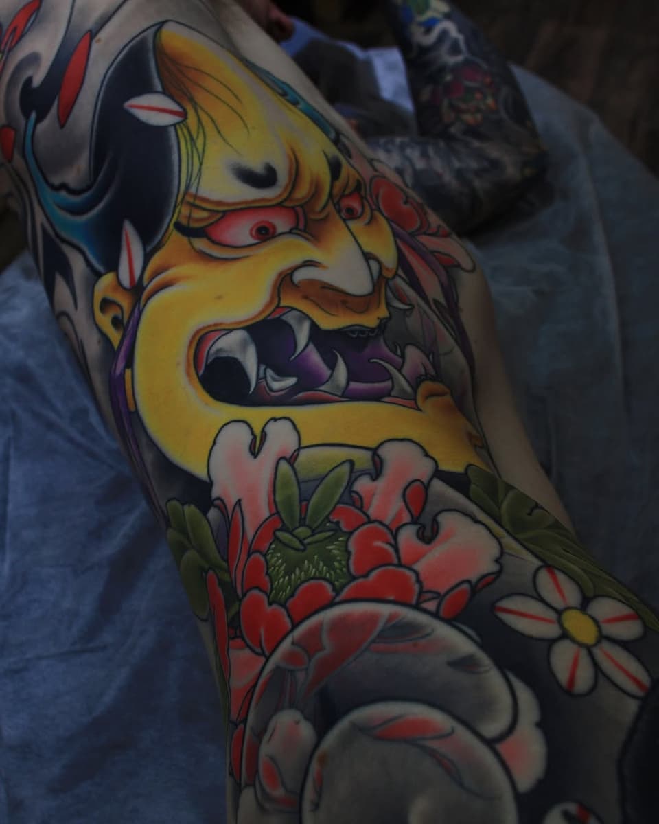 Colorful arm tattoo featuring a yellow demon face with red eyes and open mouth, surrounded by red and white flowers with green leaves.