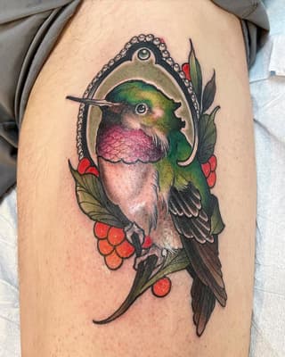 Colorful hummingbird with pink and green plumage perched on red berries and green leaves, framed in an ornamental pendant on upper arm.