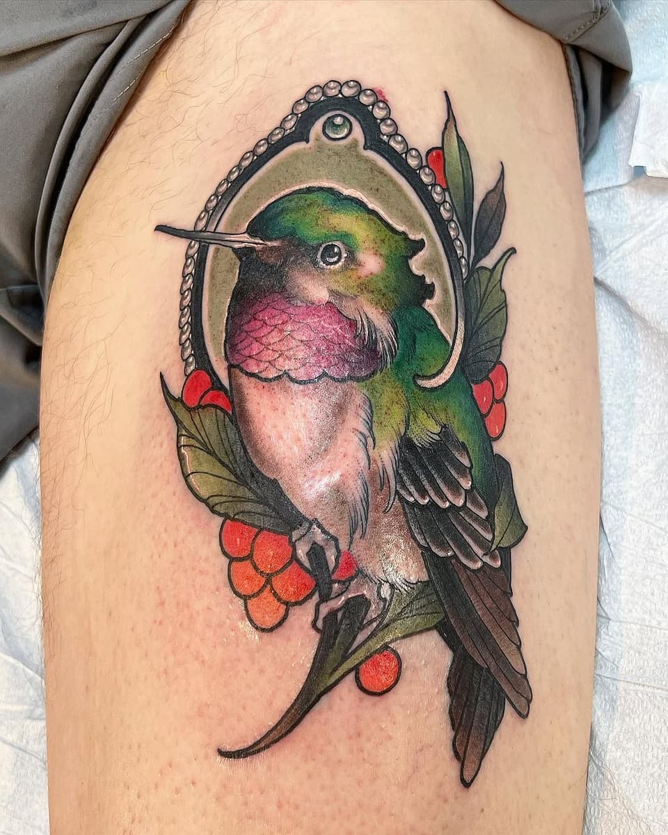 Colorful hummingbird with pink and green plumage perched on red berries and green leaves, framed in an ornamental pendant on upper arm.