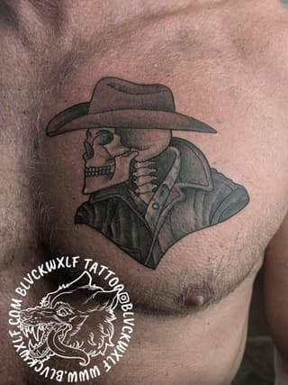 Skull wearing a cowboy hat and button-up shirt, tattooed on chest in black and gray ink, with artist watermark.