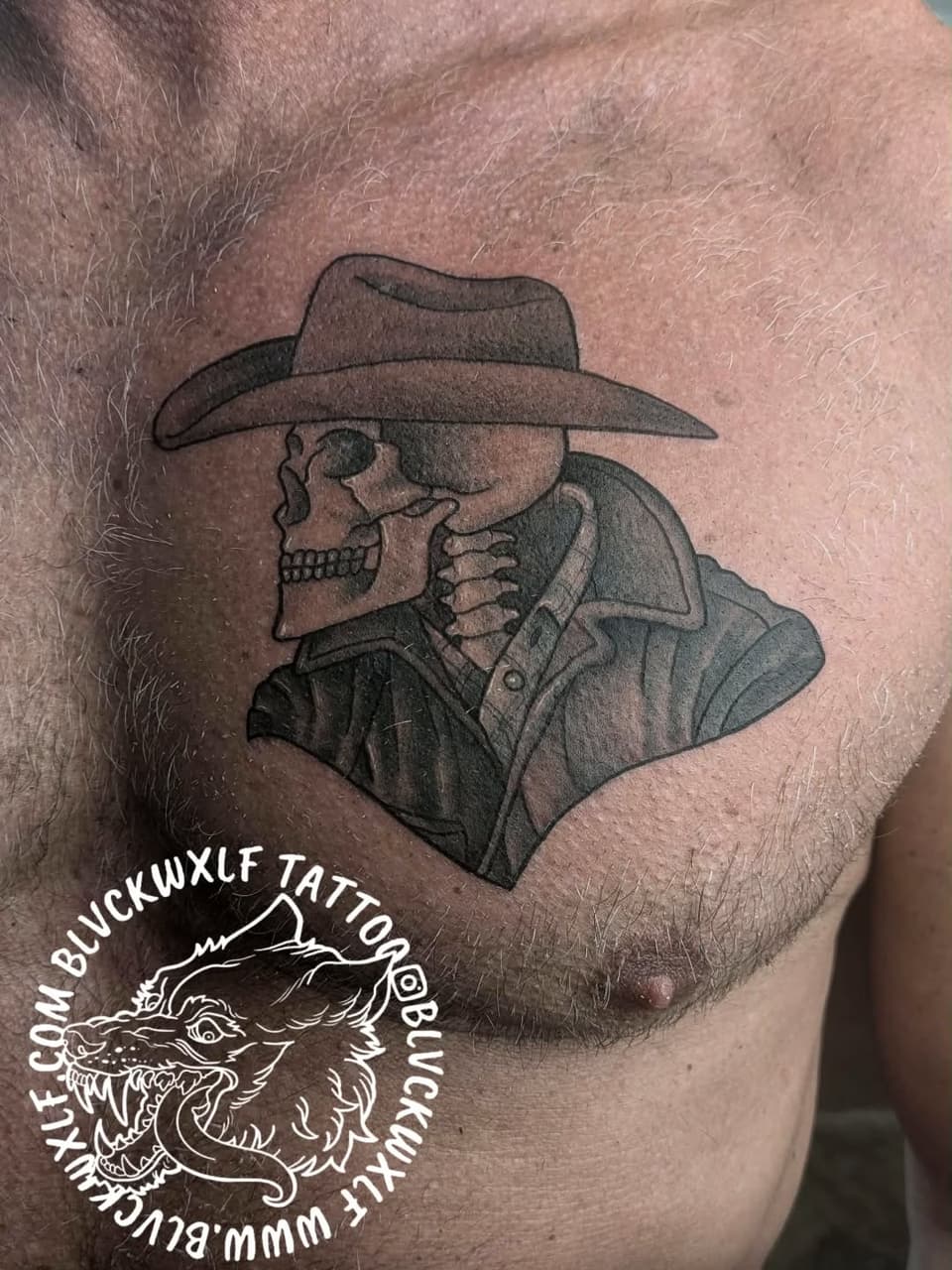 Skull wearing a cowboy hat and button-up shirt, tattooed on chest in black and gray ink, with artist watermark.