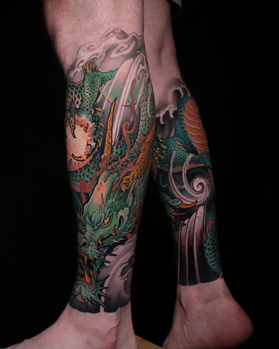 Colorful leg sleeve tattoo featuring green dragons with orange flames, ornamental scrollwork, and decorative elements wrapping around both calves.