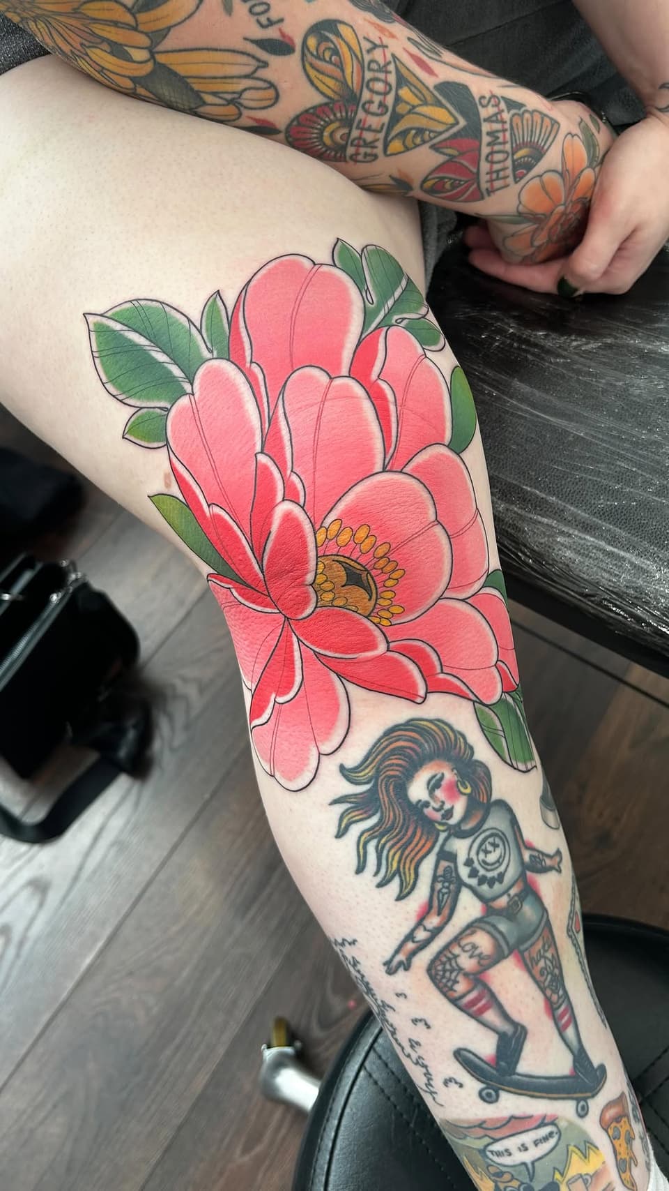 Large pink flower with yellow center and green leaves on thigh, with a female figure tattoo visible lower on the leg.