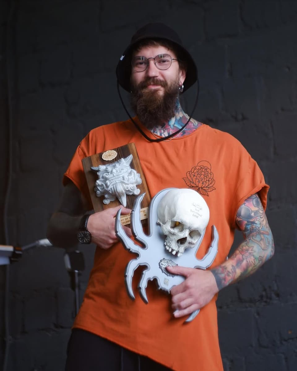 Tattooed man holding carved wooden mask and white skull sculpture, wearing orange t-shirt with skull and flower designs.