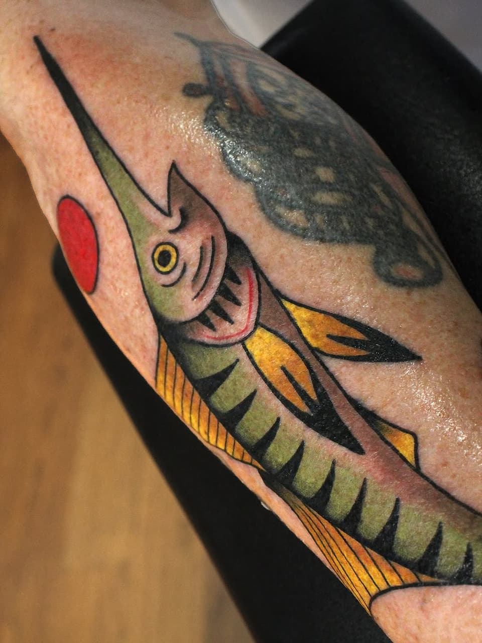 Colorful fish head tattoo on forearm featuring green, yellow, orange, and red tones with yellow eye and detailed gill markings.