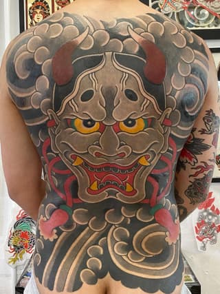 Large back tattoo featuring a beige and red demon mask with gold eyes and ornamental cloud patterns in gray and tan tones.