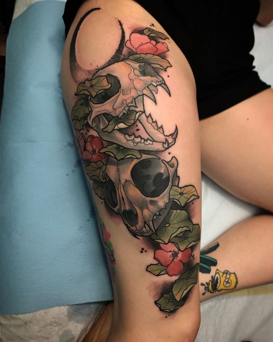 Skull with floral elements including pink flowers and green leaves wrapped around it, tattooed on the upper thigh in color.