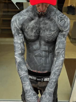 Man displays extensive gray-toned tattoos covering chest, shoulders, and both arms with skulls and ornamental designs in a portfolio photo.