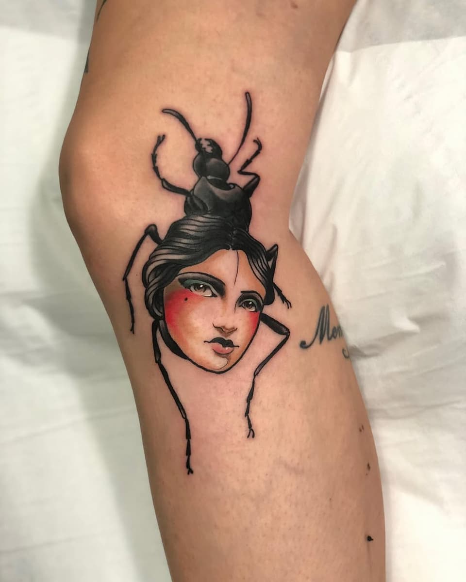 Tattoo on upper arm featuring a woman's face with red cheeks and black hair topped by a beetle, with decorative line work extending downward.