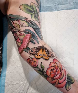 Colorful arm tattoo featuring roses, a butterfly, and botanical elements in red, orange, yellow, and green on the forearm and upper arm.