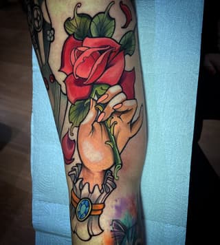 Hand holding a red rose with green leaves and stem, tattooed on the leg in red, green, orange, and skin tones with a jeweled bracelet detail.