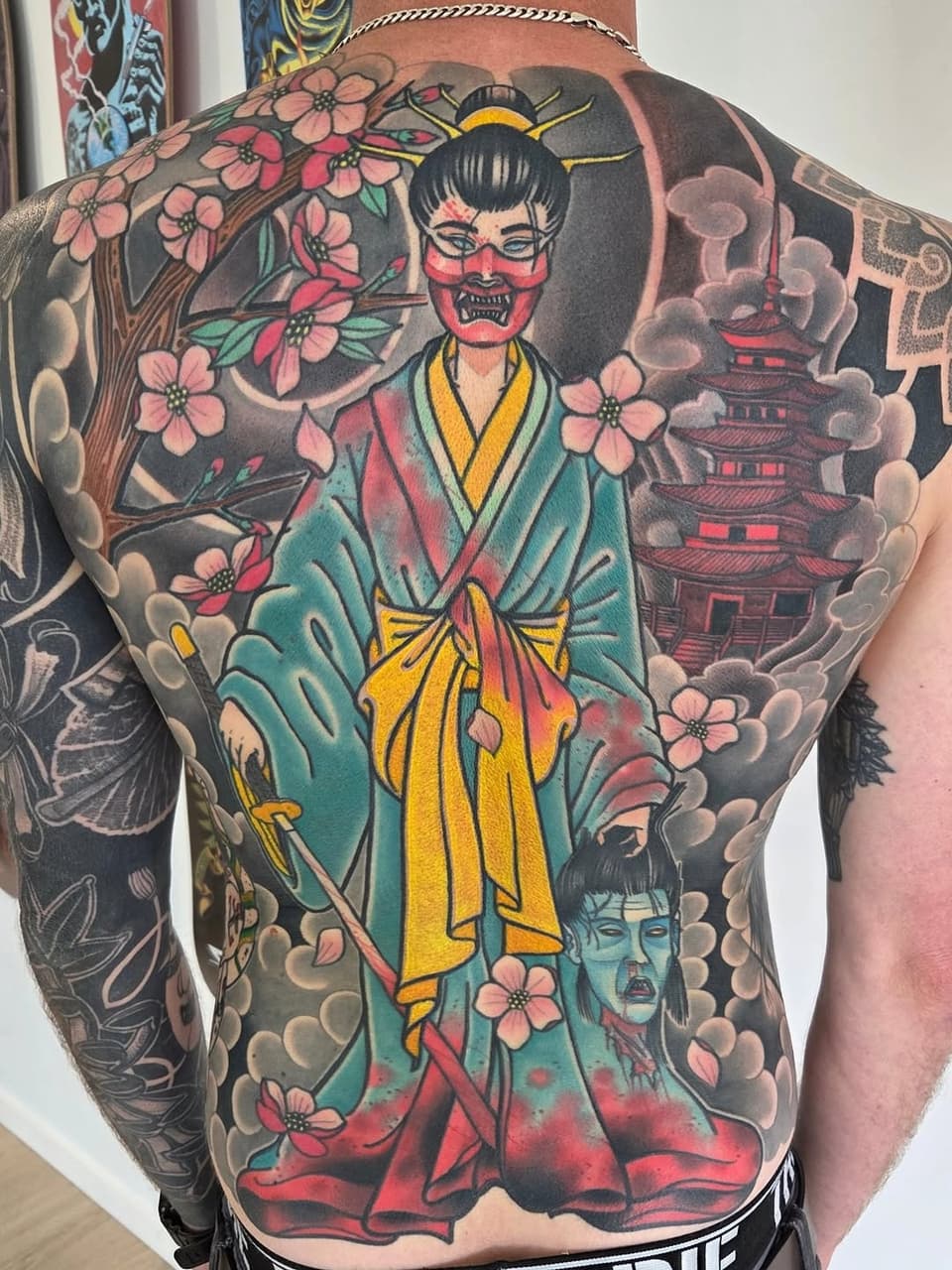 Large back tattoo featuring a demon-faced figure in teal and yellow robes surrounded by pink flowers, a pagoda, clouds, and a smaller demon head below.