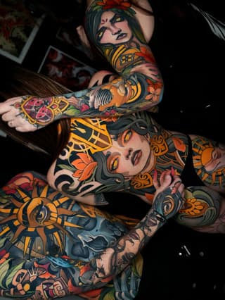 Two women with full-body colorful tattoos featuring portrait faces, flowers, ornamental patterns, and decorative elements in orange, red, yellow, and green.