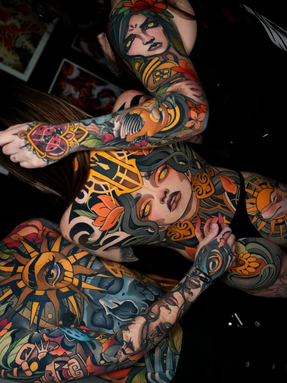 Two women with full-body colorful tattoos featuring portrait faces, flowers, ornamental patterns, and decorative elements in orange, red, yellow, and green.
