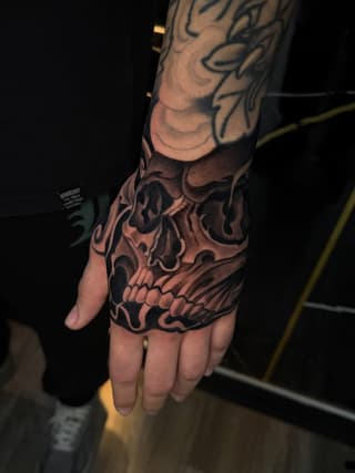 Detailed skull tattoo covering the back of a hand and extending up the forearm, rendered in black and brown ink with shading and ornamental details.