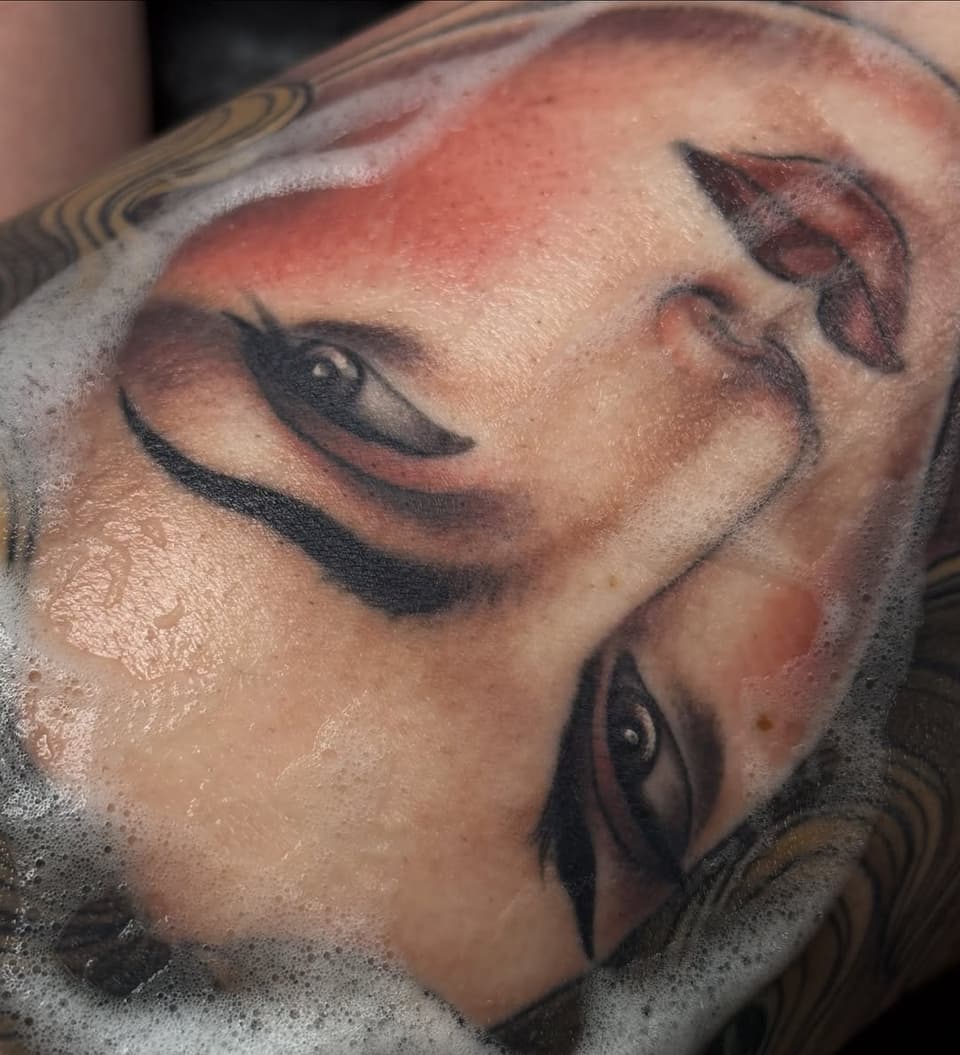Detailed portrait tattoo on upper arm showing a man's face with closed eyes and facial hair, rendered in black and brown ink with shading.