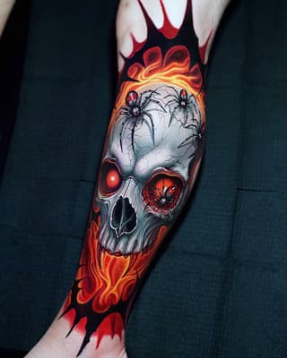 Skull tattoo with spiders, glowing red eyes, and orange-yellow flames on the forearm against black clothing.