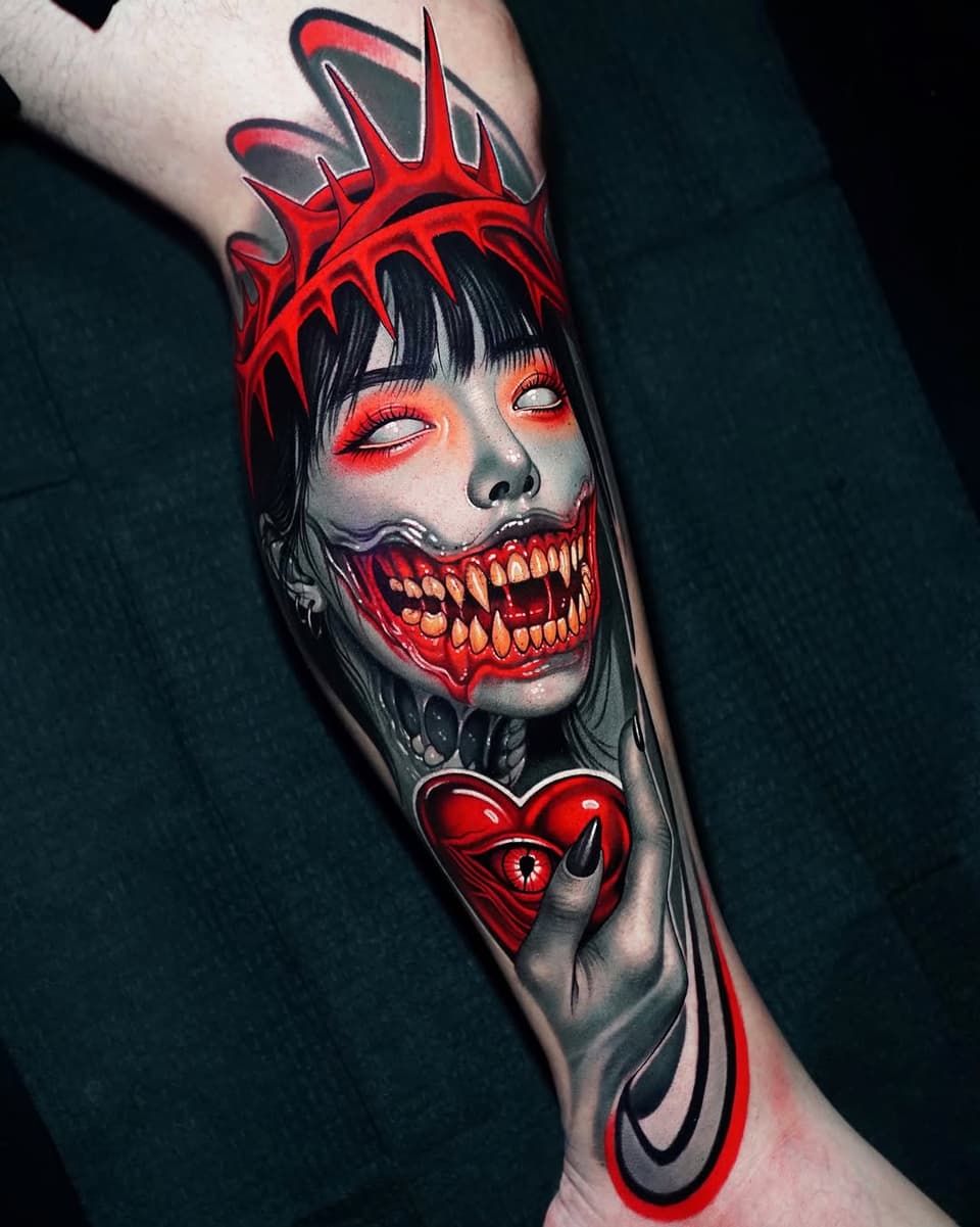 Colorful arm tattoo featuring a demonic female face with red spiky crown, glowing eyes, sharp teeth, and a red heart with an eye below the neck.