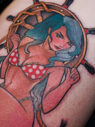 Colorful tattoo of a smiling woman with teal hair wearing a red polka dot bikini top and steering wheel, on upper arm.