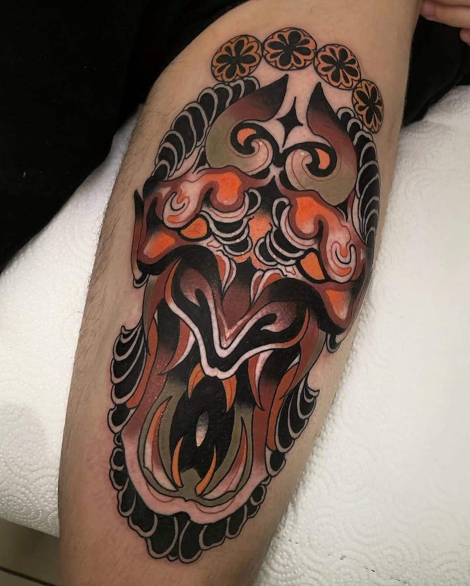 Colorful demon or oni mask tattoo on the calf featuring orange, red, and black tones with ornamental floral border details.