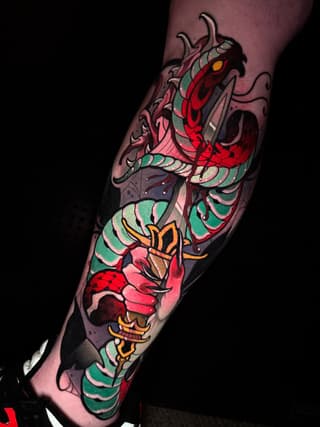 Colorful tattoo on a forearm featuring a demon head with horns and glowing eyes, layered with red flowers and green scaled wings in vibrant reds, greens, oranges, and pinks.