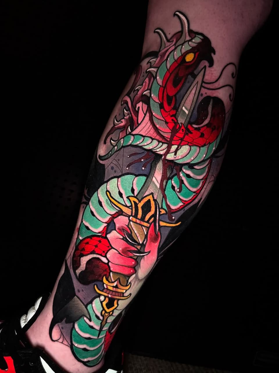 Colorful tattoo on a forearm featuring a demon head with horns and glowing eyes, layered with red flowers and green scaled wings in vibrant reds, greens, oranges, and pinks.