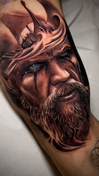 Detailed portrait tattoo of a bearded man's face with blue eyes and a small flying insect on his forehead, rendered in brown and tan tones on the arm.