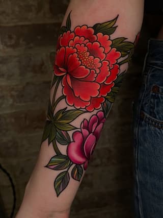 Red and pink layered flowers with olive-green leaves tattooed on the forearm, featuring detailed petals and a central stamen in warm tones.