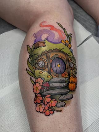 Colorful tattoo on a calf depicting a hobbit hole with a round purple door, surrounded by flowers, mushrooms, moss, and a purple swirling sky above.