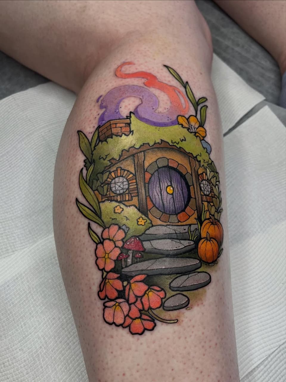 Colorful tattoo on a calf depicting a hobbit hole with a round purple door, surrounded by flowers, mushrooms, moss, and a purple swirling sky above.