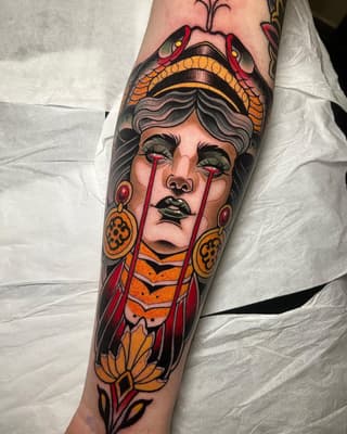 Colorful tattoo on forearm featuring a woman's face with tears, ornamental headdress, dangling earrings, and flowering vines in red, orange, and gold tones.