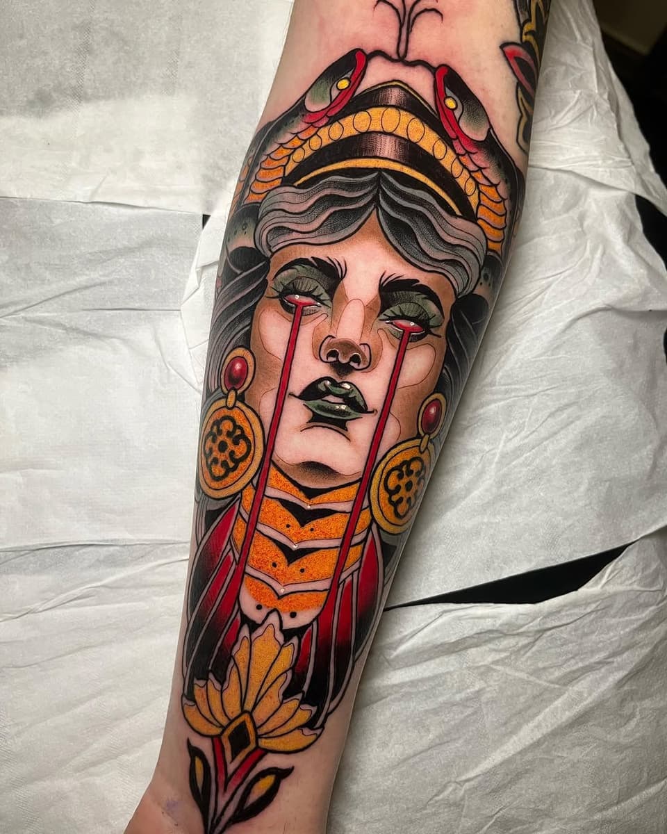 Colorful tattoo on forearm featuring a woman's face with tears, ornamental headdress, dangling earrings, and flowering vines in red, orange, and gold tones.