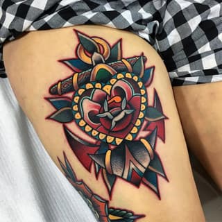 Colorful heart tattoo with ornamental beading and leafy flourishes in red, gold, teal, and black on the thigh.