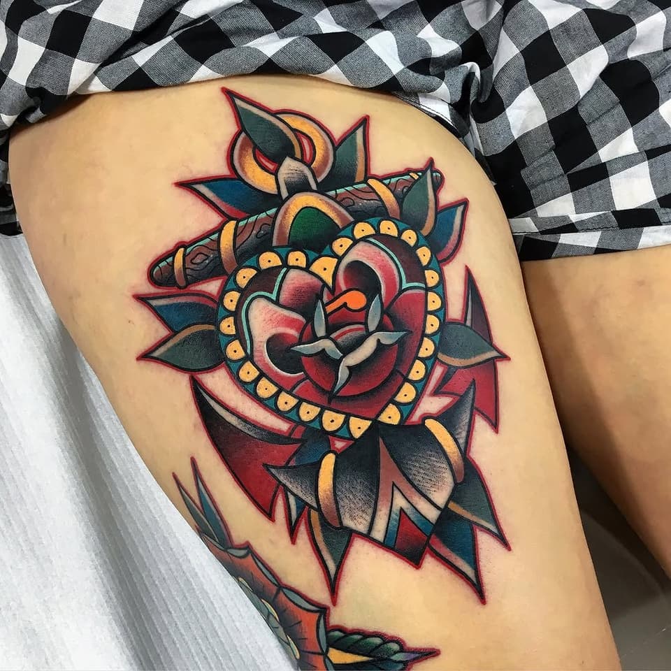 Colorful heart tattoo with ornamental beading and leafy flourishes in red, gold, teal, and black on the thigh.