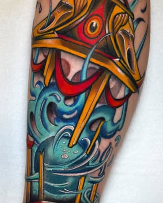 Colorful leg tattoo featuring stacked ornamental masks in red, gold, teal, and black with decorative swirling elements and striped accents.