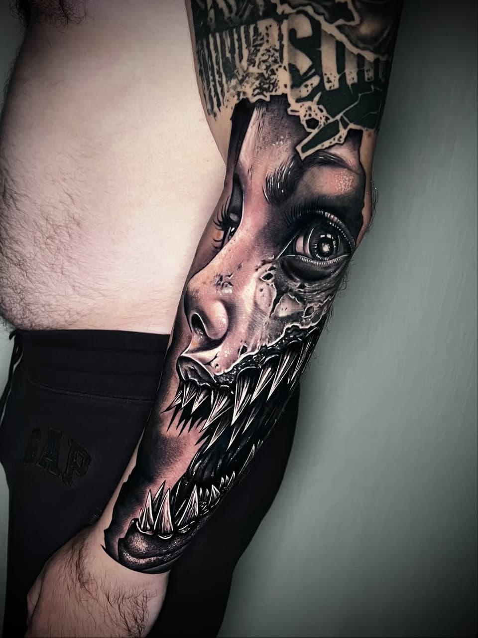Detailed demon face tattoo on upper arm with sharp teeth, piercing eye, and monochromatic shading in black and gray tones.