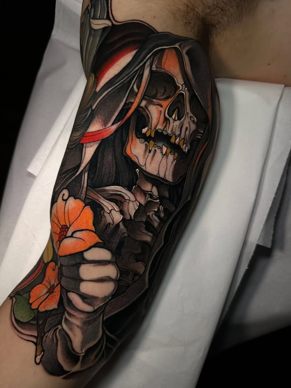 Colorful skull tattoo with long hair, red ribbon, and orange flowers on a person's forearm and upper arm.