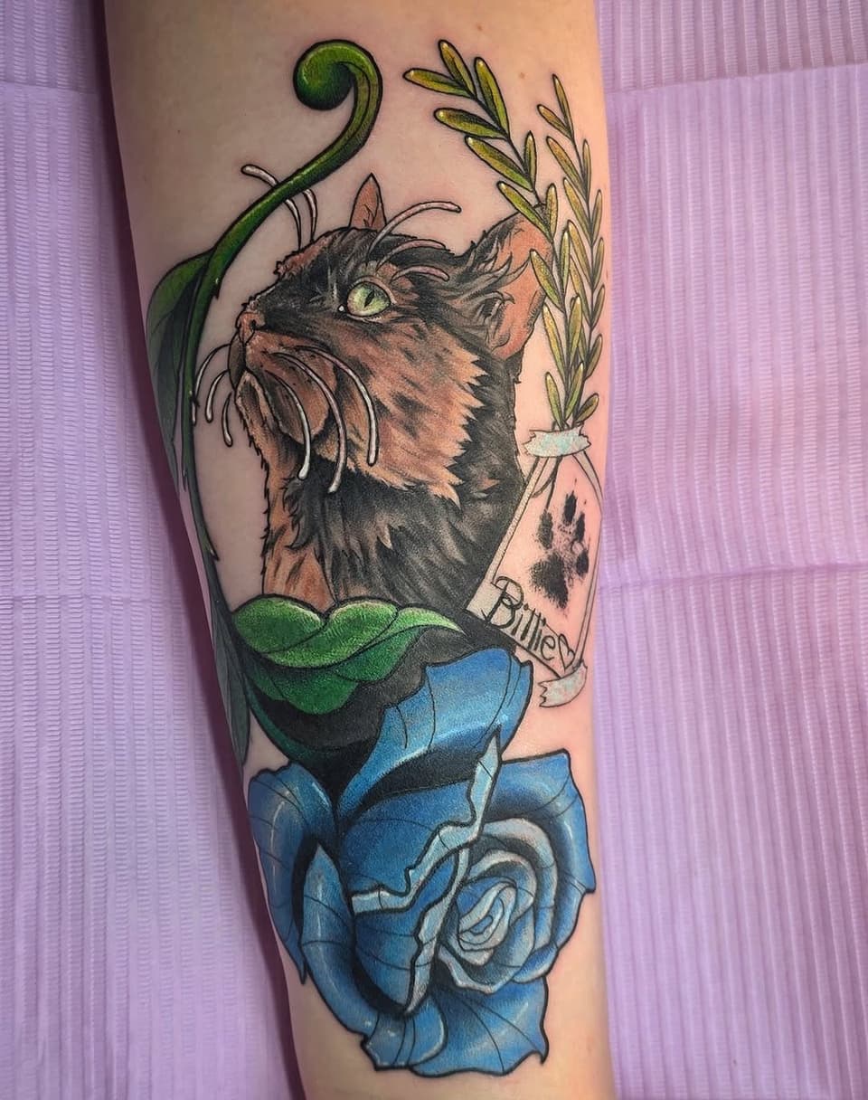 Colorful cat tattoo on forearm featuring a tabby cat with green vine and leaf elements, blue roses, and text reading "Blurred," displayed on pale skin.