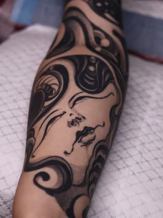 Tattoo on forearm featuring a profile face surrounded by ornamental swirling designs in black ink on skin.