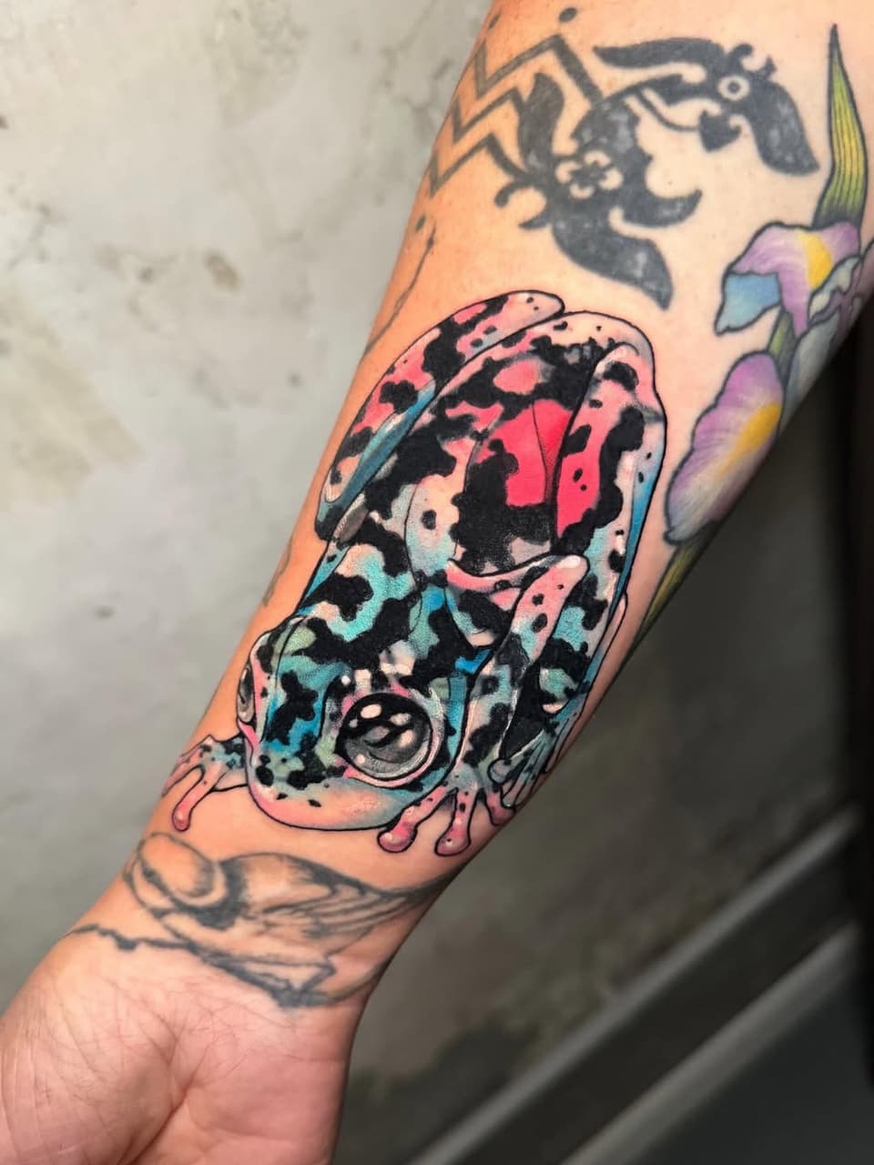 Colorful spotted frog or toad tattoo on forearm with pink, blue, and black patches, surrounded by smaller black silhouette designs on the arm.
