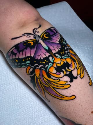 Colorful butterfly tattoo with purple, orange, and yellow wings on a person's forearm, featuring detailed wing patterns and decorative antennae.