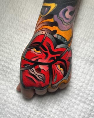 Red demonic mask face tattoo with yellow eyes and orange horns on hand, featuring purple and yellow elements on upper forearm.
