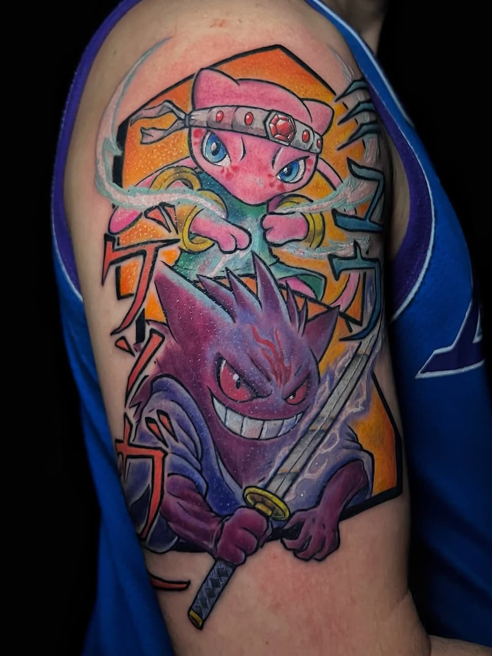 Colorful shoulder tattoo featuring a pink character with a headband and a purple demon face below, holding a sword, with orange and yellow background elements.