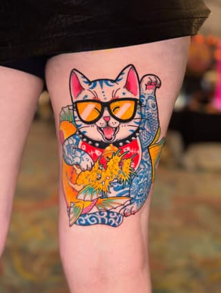 Colorful Maneki-neko cat wearing sunglasses with raised paw and koi fish details tattooed on thigh in orange, blue, red, and pink.