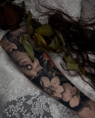 Arm tattoo featuring floral and botanical elements in black and gray ink, displayed on light fabric with dried flowers.