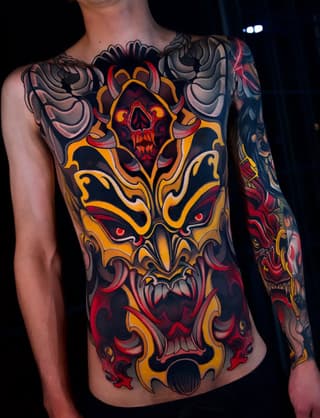 Colorful chest and sleeve tattoo featuring stacked demon faces with skull imagery, ornate gold scrollwork, and red accents on bare-chested male torso.