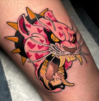 Pink demon or oni head with red hearts, gold spikes, and yellow eyes tattooed on an upper arm in red, pink, black, and gold colors.
